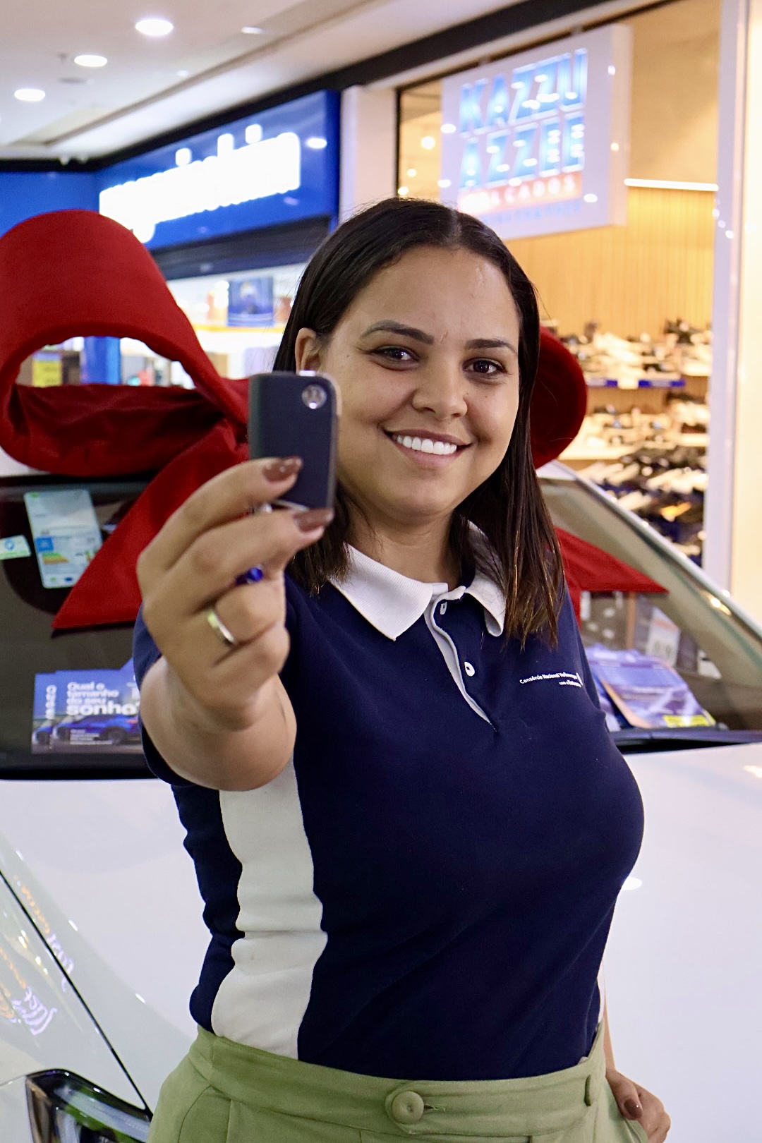 Aline Assis sorrindo em frente a um carro, segurando um celular