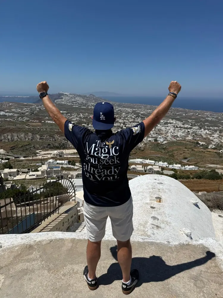 Magic with his arms raised above Santorini, the back of his navy shirt reading The Magic you seek is already in you